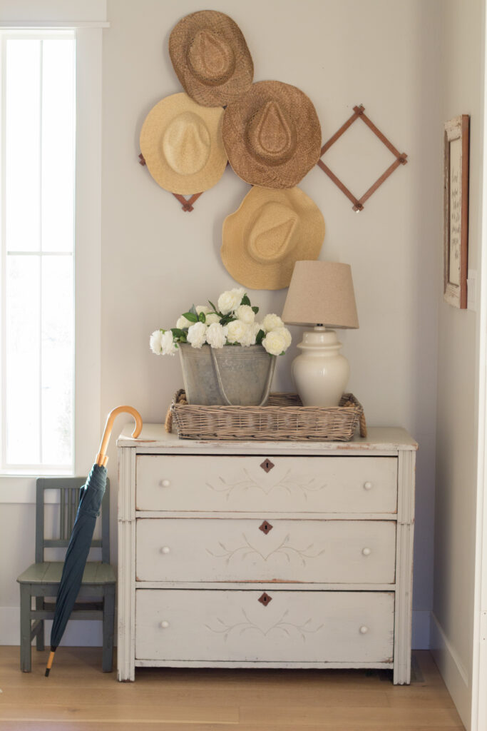 a spring decorating idea for a 3 drawer dresser - a basket is on top of it with a bucket of flowers and lamp in it. Above the dresser is an arrangment of 4 straw hats and an umbrella is leaning against the dresser.