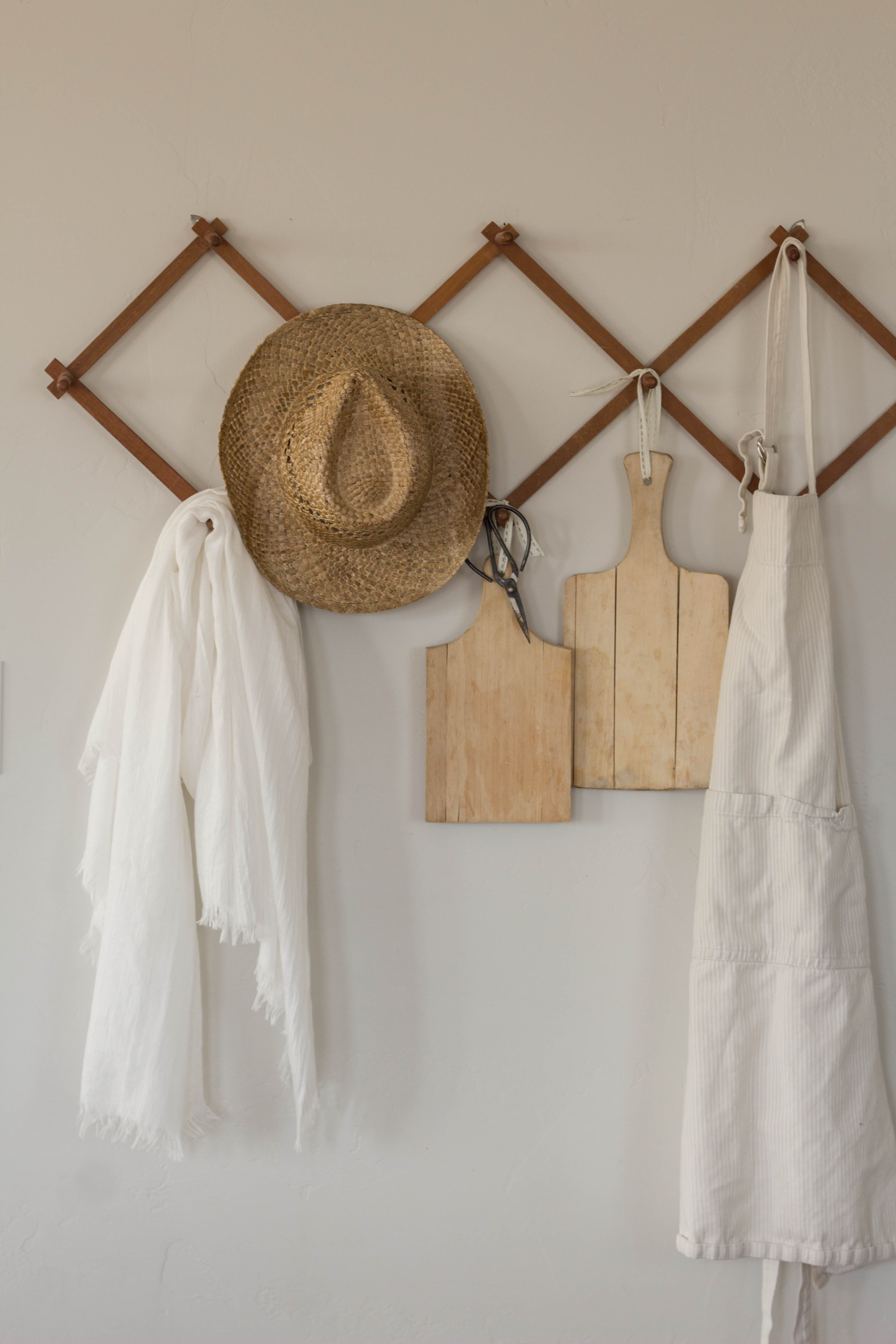 a hanging peg rail on the wall with a straw hat, floral shears, cutting boards, white apron and white scarf hanging from the pegs. It's a cheery and bright early spring decorating idea.