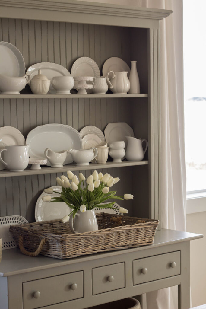 a gray hutch filled with white dishes and a basket with an arrangment of faux tulips in it