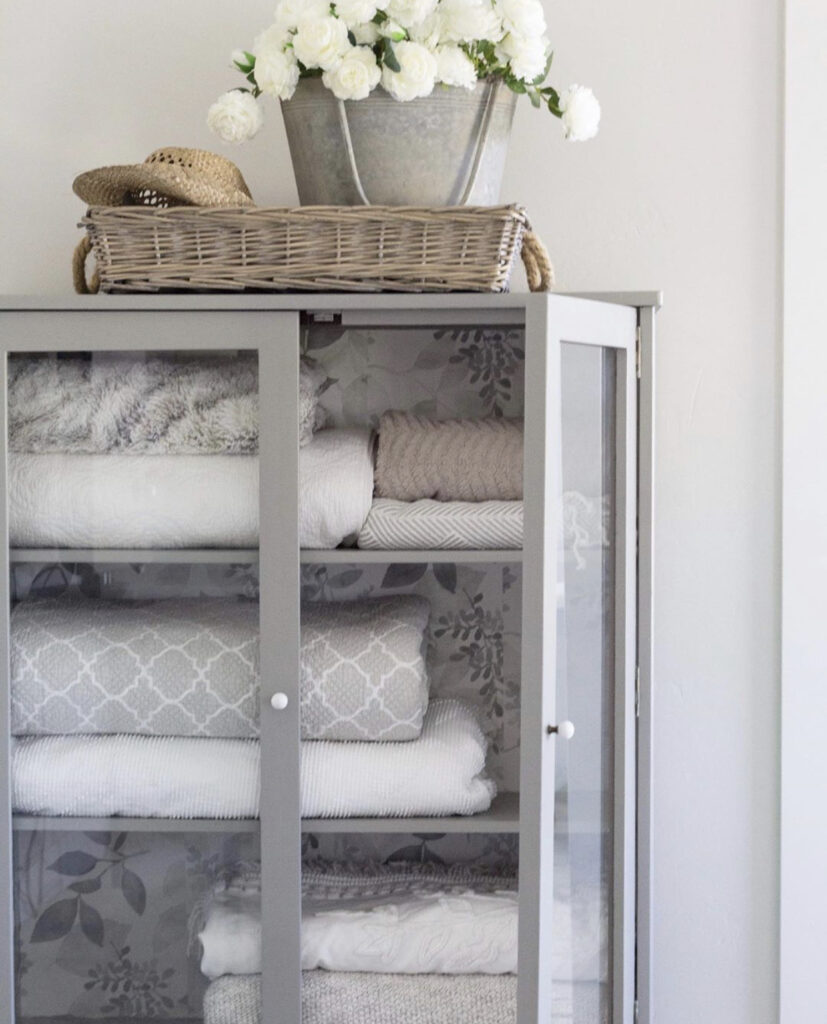 A linen cupboard with folded blankets and the same basket with the bucket of flowers in it that was in the mudroom and on top of the dresser.